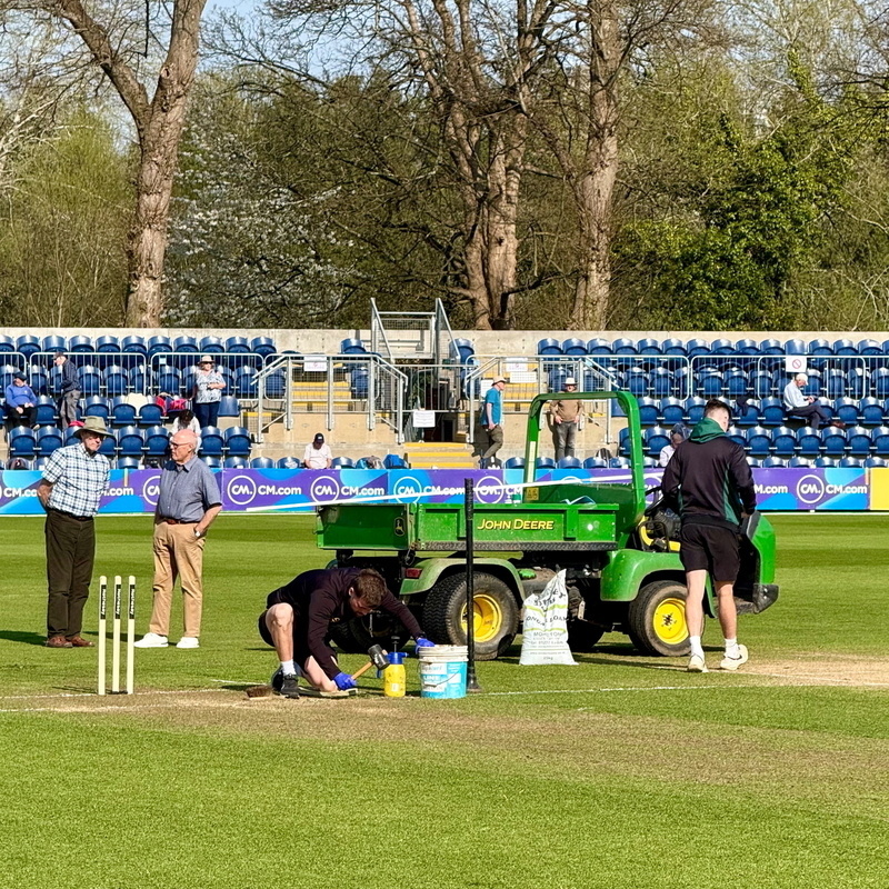 Auto-generated description: A group of people are preparing a cricket pitch with equipment, including a small green vehicle, while others watch from the stands.