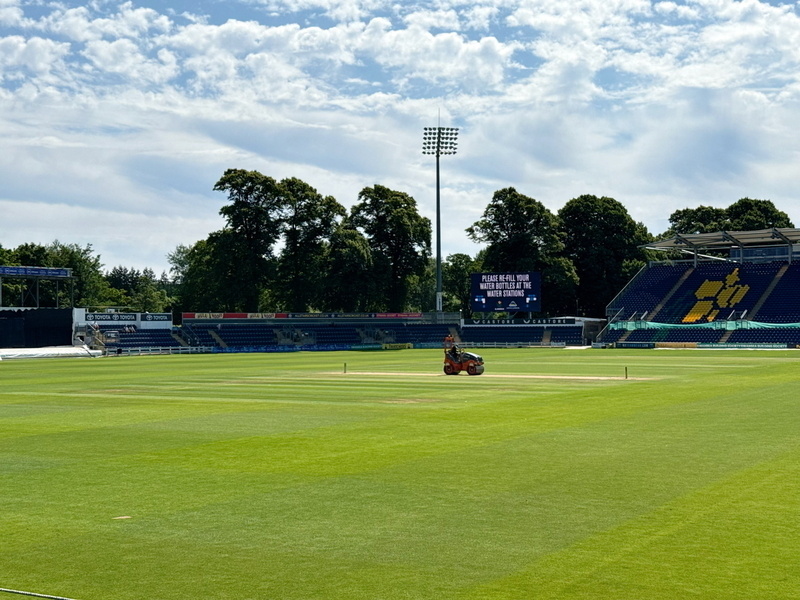 Auto-generated description: A person rides a lawnmower on a cricket field under a partly cloudy sky.
