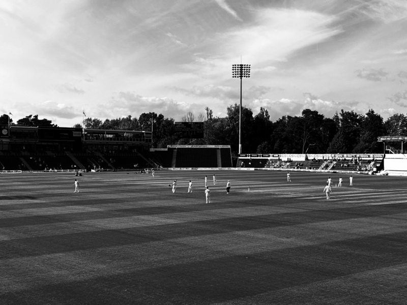 Auto-generated description: A black and white photo shows a cricket match in progress with spectators and trees in the background.