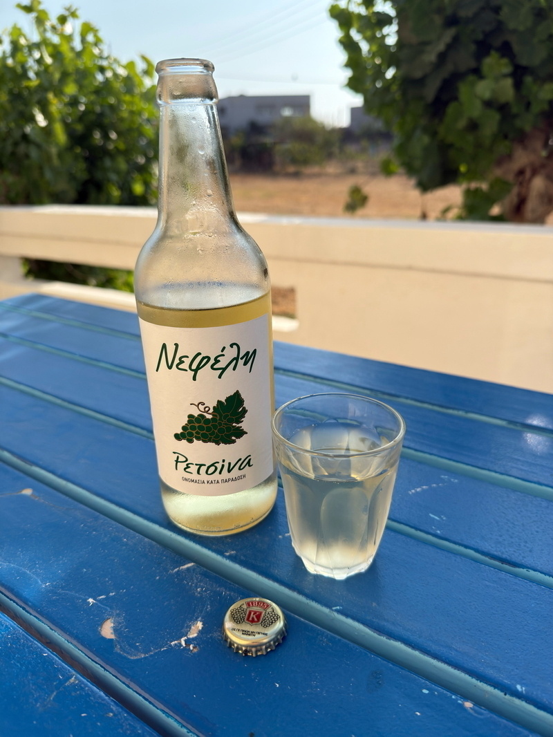 Auto-generated description: A bottle of retsina wine and a glass sit on a blue table with a bottle cap nearby, against a background of greenery and a sunlit landscape.