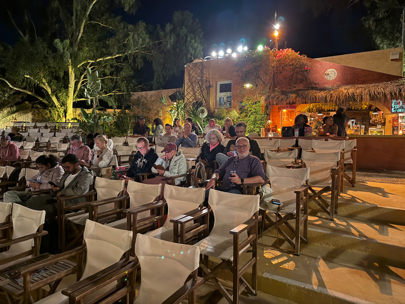 Auto-generated description: A group of people is seated in an outdoor venue at night, with a few individuals standing near a brightly lit bar in the background.