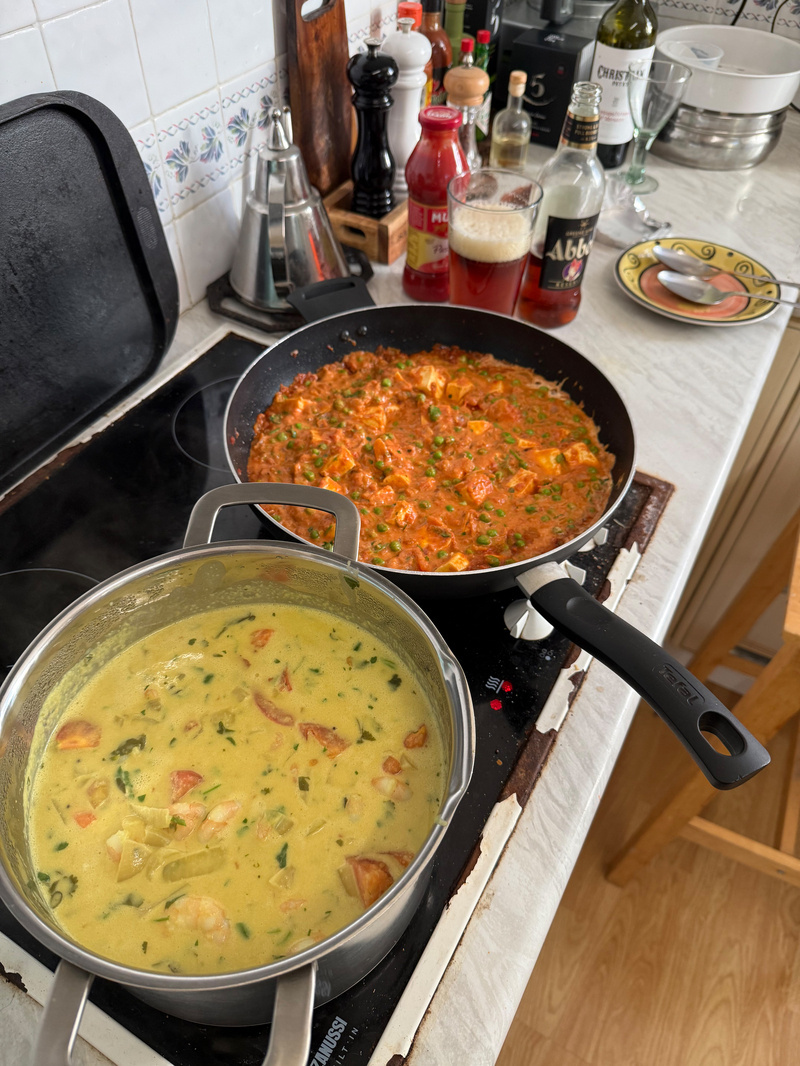 Auto-generated description: Two pans on a stove, one with a yellow prawn curry and the other with a red paneer curry, are surrounded by various condiments in a kitchen setting.