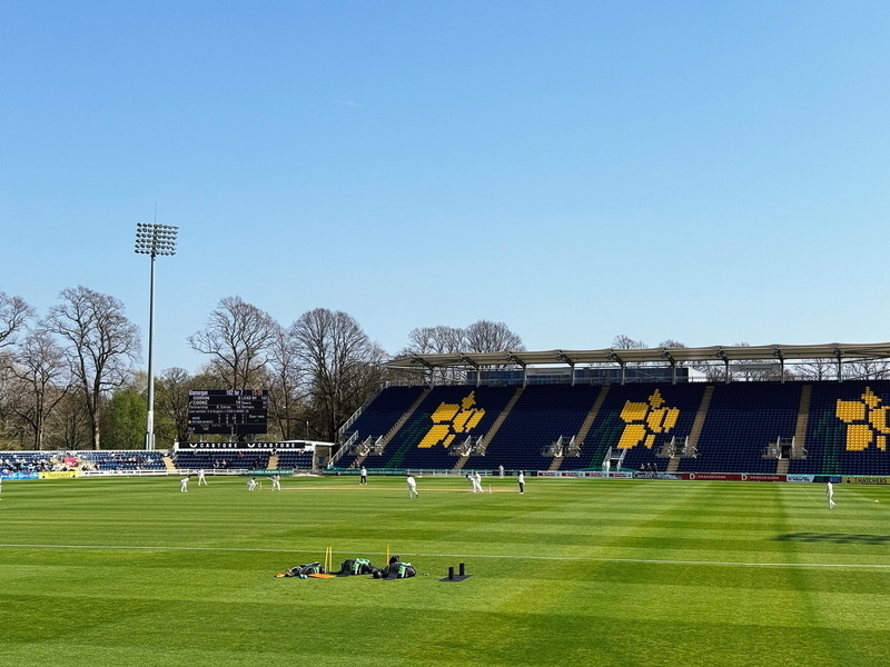 Auto-generated description: A cricket match is taking place on a sunny day in a stadium with blue seating and a partially filled crowd.
