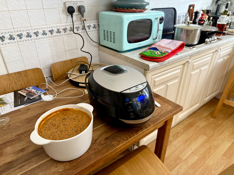 Auto-generated description: A kitchen scene features a wooden table with a pot of dal and a rice cooker, set against a background of a microwave, pots, and cooking utensils on the counter.