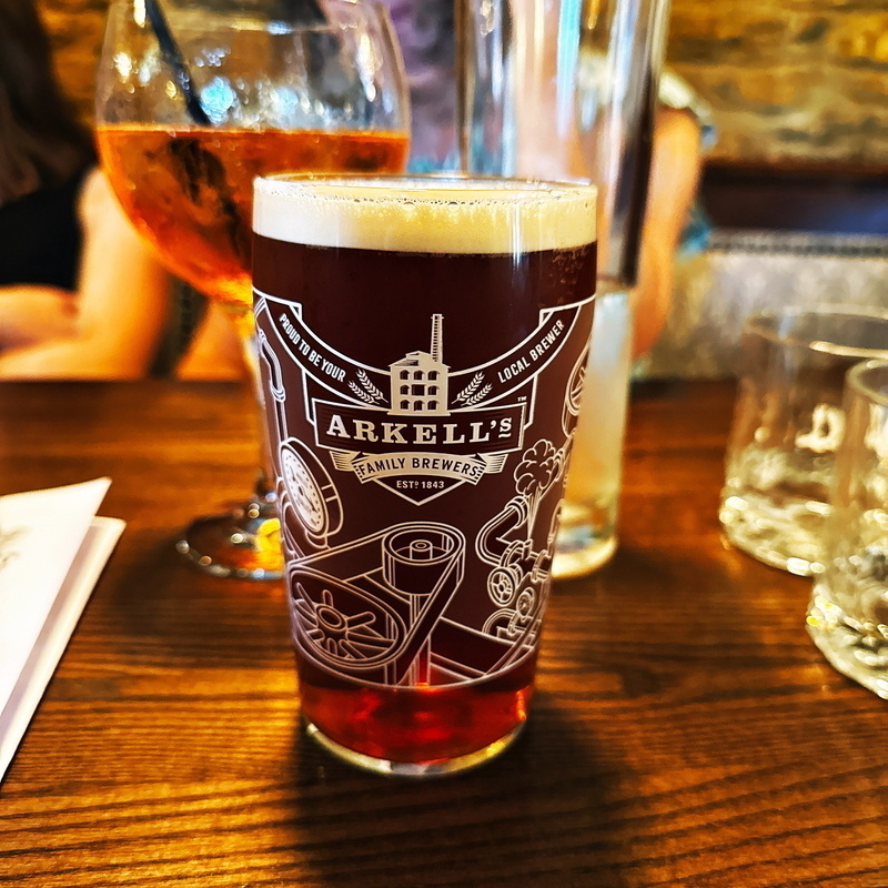 A beer glass with the label Arkell's Family Brewers containing a dark beer, placed on a wooden table. In the background, there's a larger glass with a straw and some additional glassware.