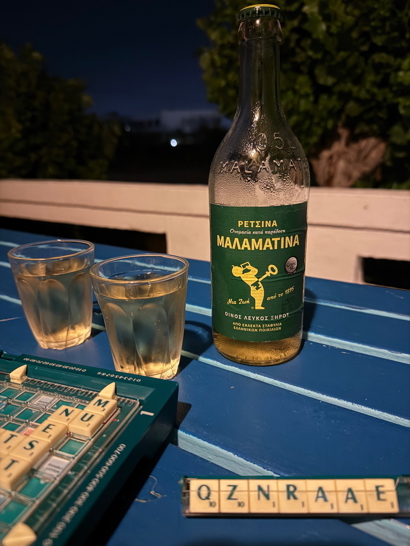 Auto-generated description: A bottle of Malamatina and two glasses of wine sit on a blue table next to a Scrabble game in progress at night.