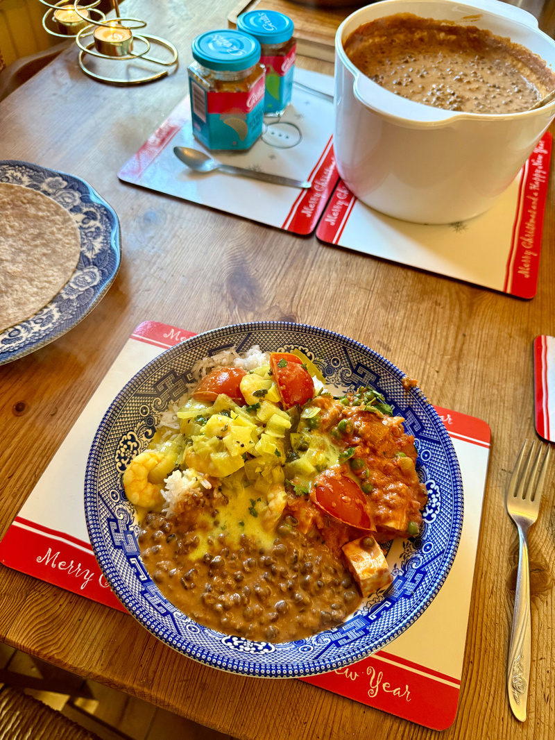 Auto-generated description: A bowl filled with a colorful assortment of foods, including lentils, vegetables, and a creamy sauce, is placed on a festive table setting with flatbreads, chutneys, and a large pot of dal in the background.