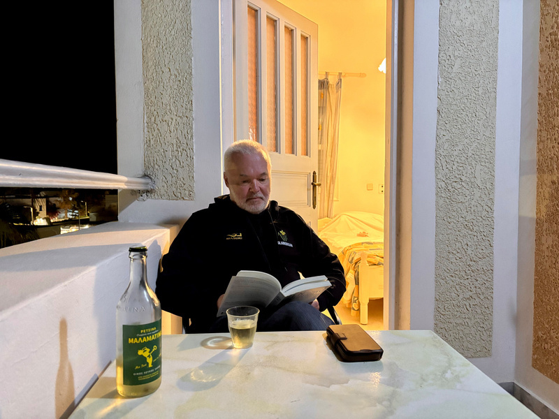 Auto-generated description: A man sits on a terrace, reading a book with a bottle and glass on the table, next to a warmly lit room.