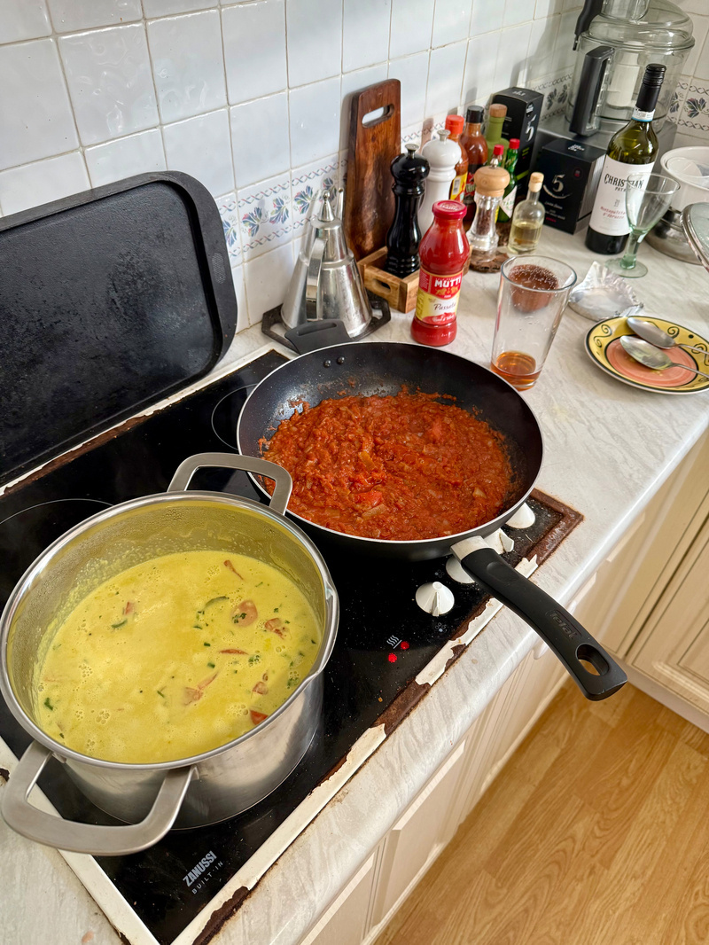 Auto-generated description: A stovetop holds two pans, one with a creamy coconut dish and the other with a tomato-based sauce, surrounded by various condiments and a bottle of wine.