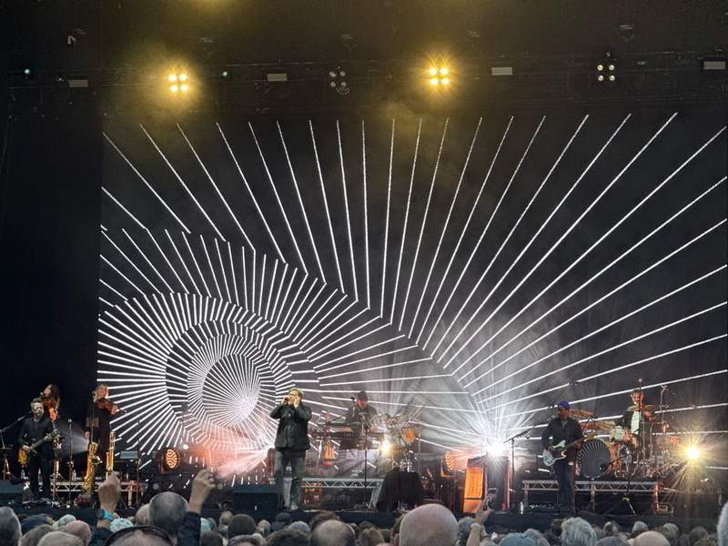 Auto-generated description: A band performs on stage with a striking black and white geometric pattern displayed on a large screen behind them.