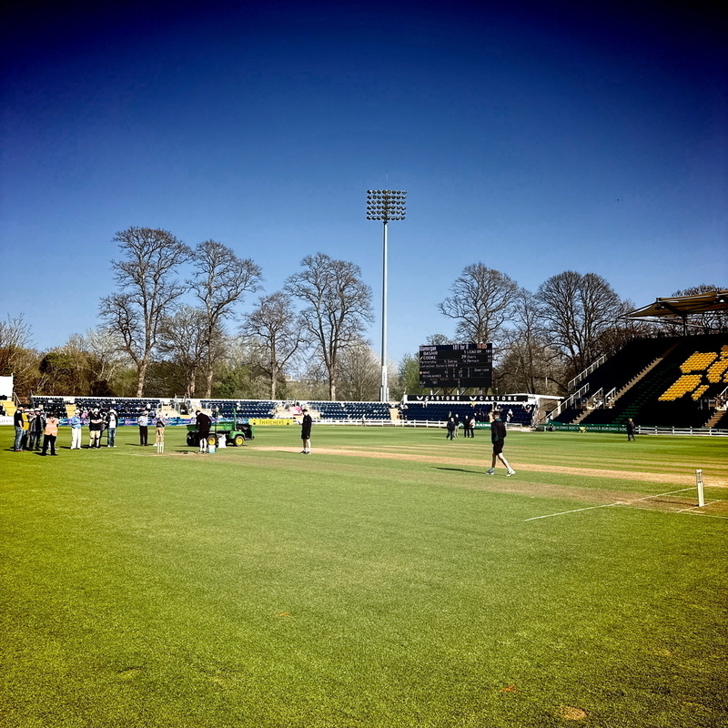 Auto-generated description: A group of people are gathered on a green cricket field under a clear blue sky.