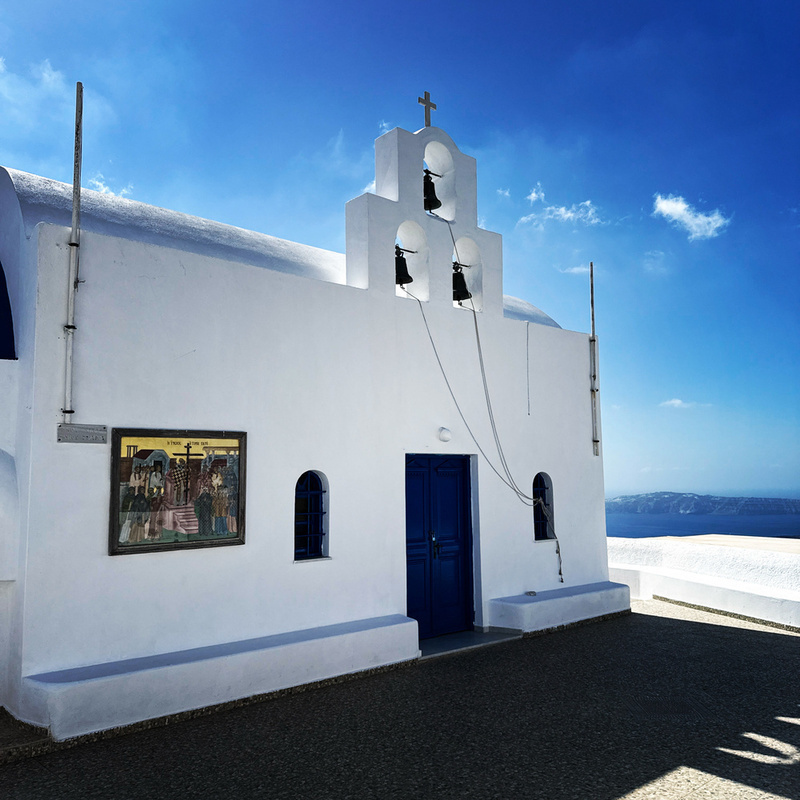 A whitewashed church with blue accents and bell towers is set against a clear blue sky and sea.