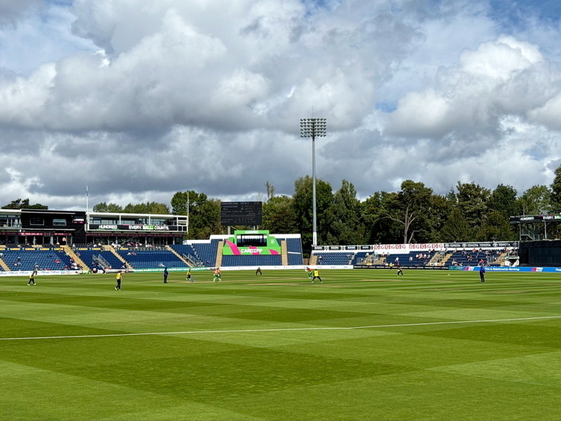 A cricket match is taking place in a stadium with a few players on the green field. The stands are partially filled, and the scoreboard is visible. The sky is cloudy with patches of blue.