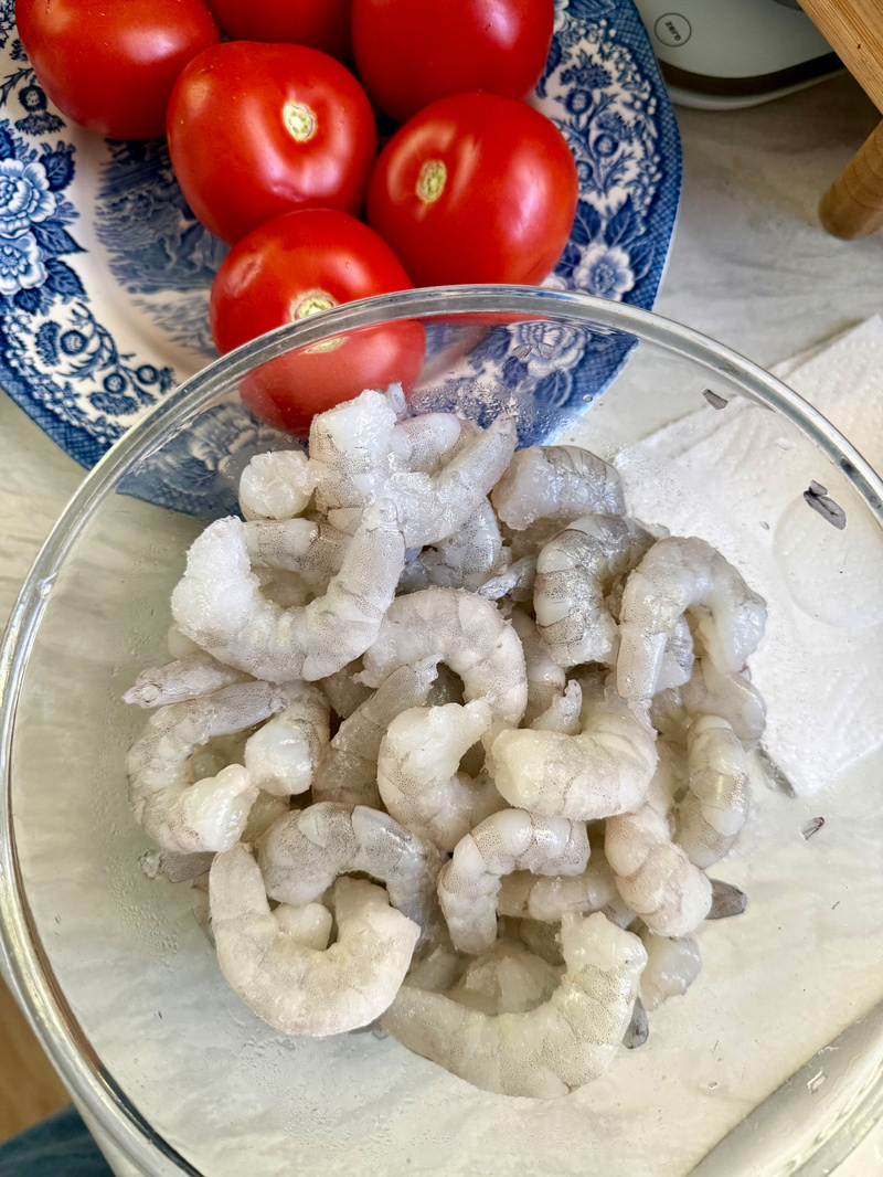 Auto-generated description: A bowl of raw prawns is placed next to a plate with red tomatoes.