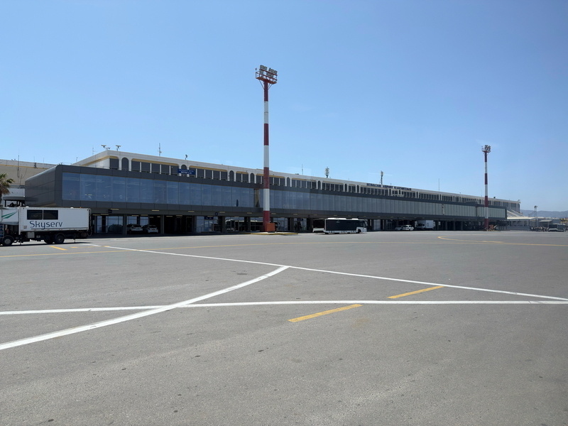 Auto-generated description: A large, modern airport terminal with several vehicles in front and clear blue skies above.
