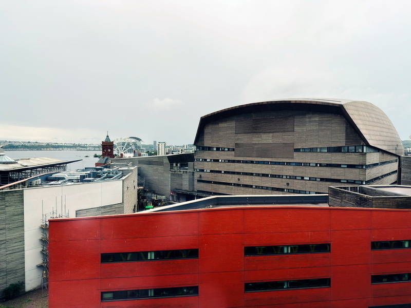The image shows a large modern building with a distinctive, curved roof, likely an arts or cultural center, set against a cloudy sky. In the foreground, there is a red brick building. In the background, a river with a bridge and Ferris wheel.