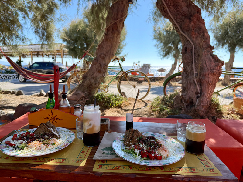 Auto-generated description: Two plates of food and drinks are set on a colorful table with a view of trees and the sea in the background.
