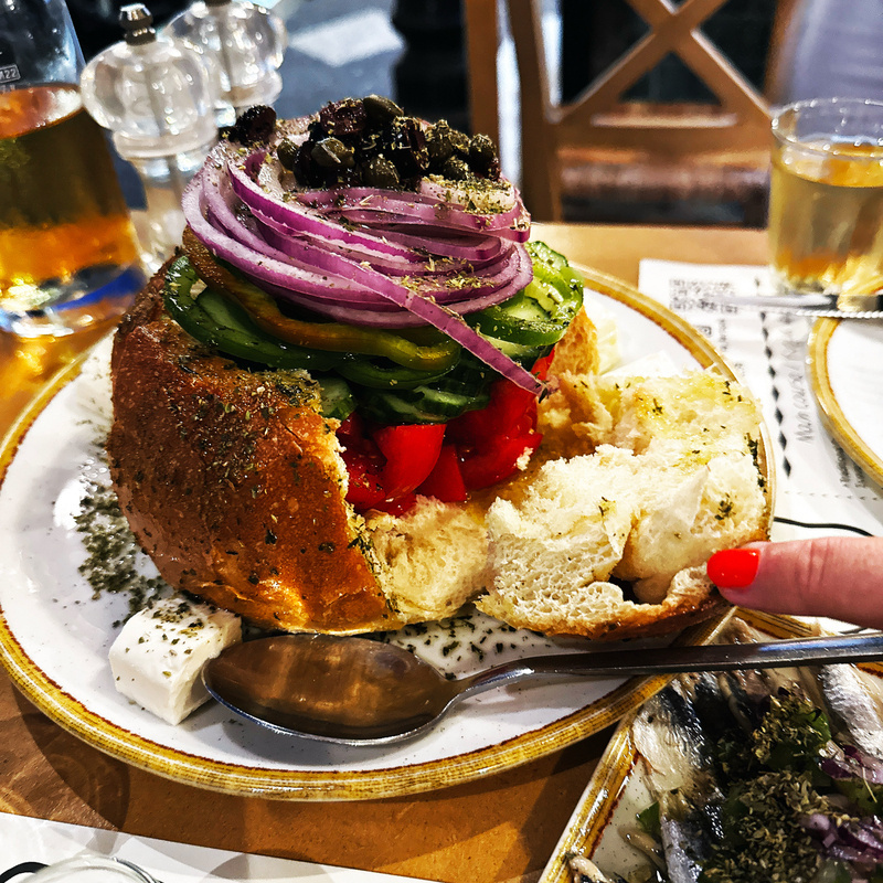 A hollowed bread bowl is filled with a colorful assortment of vegetables, including slices of cucumber, tomato, red onion, and olives, and topped with herbs.