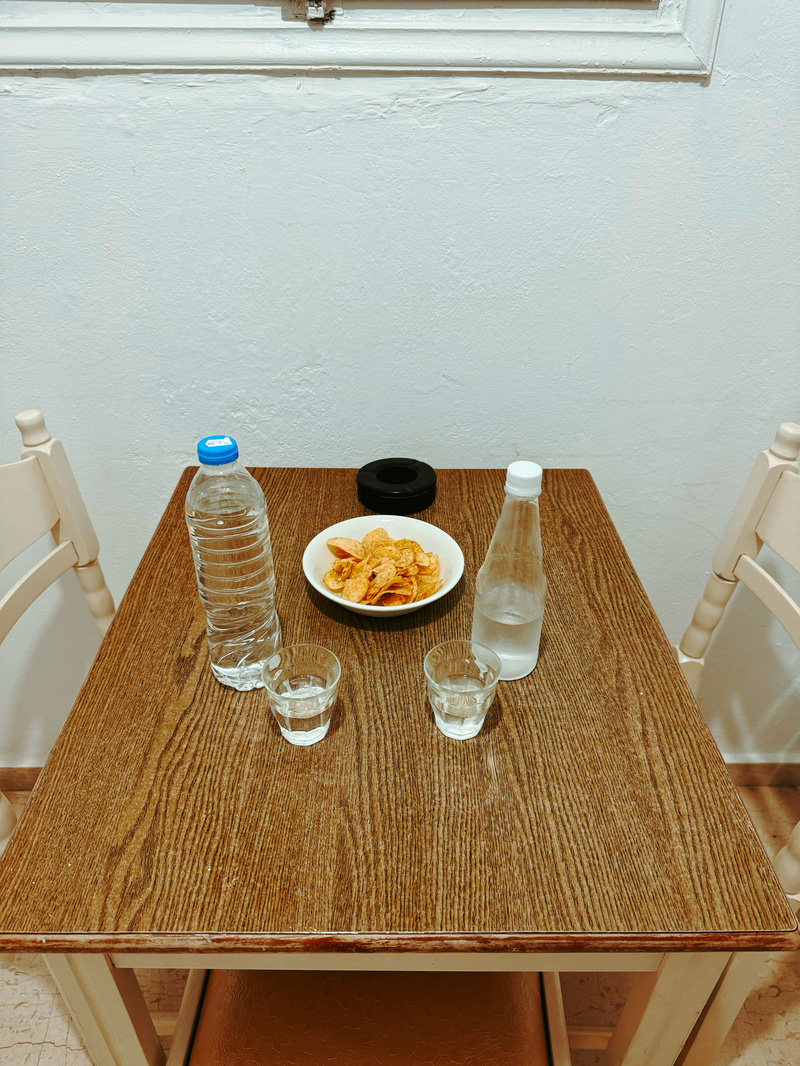 A small wooden table set for two features a bowl of crisps, two bottles of clear liquid, and two glasses.