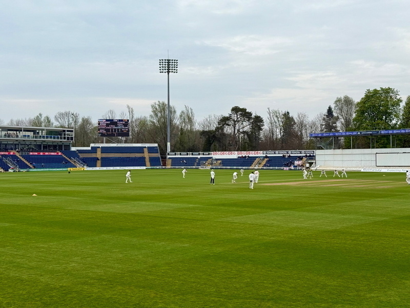 Auto-generated description: A cricket match is taking place on a green field with players dressed in white.