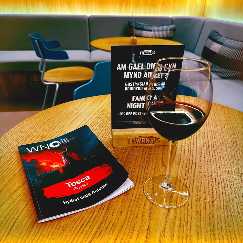 A table with a glass of red wine, a brochure for Tosca by Puccini, and a sign offering a 10% discount on drinks post-show. The setting appears to be a cozy lounge with modern seating in the background.