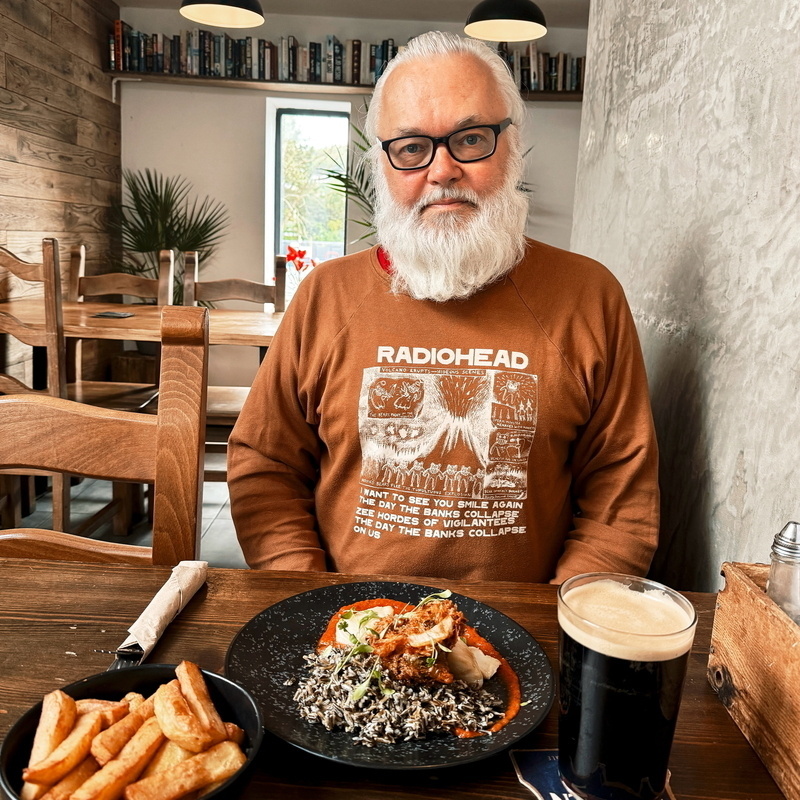 Auto-generated description: A man with a white beard is sitting at a table in a cozy restaurant, wearing a Radiohead sweatshirt and enjoying a meal with a glass of dark beer and a side of fries.