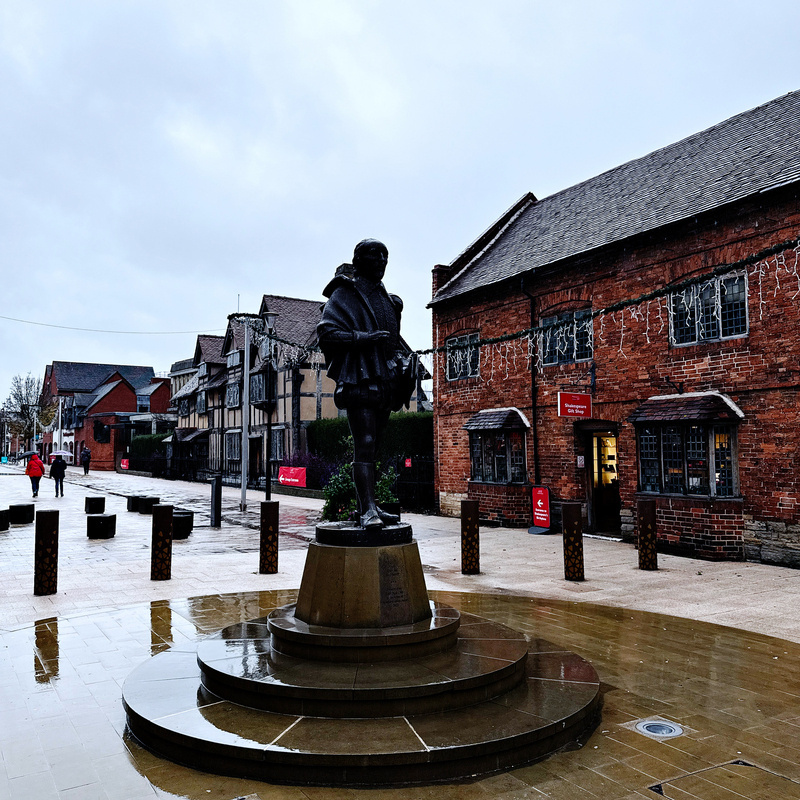 Auto-generated description: A statue stands on a pedestal in a paved outdoor area surrounded by historic brick buildings and festive lights.