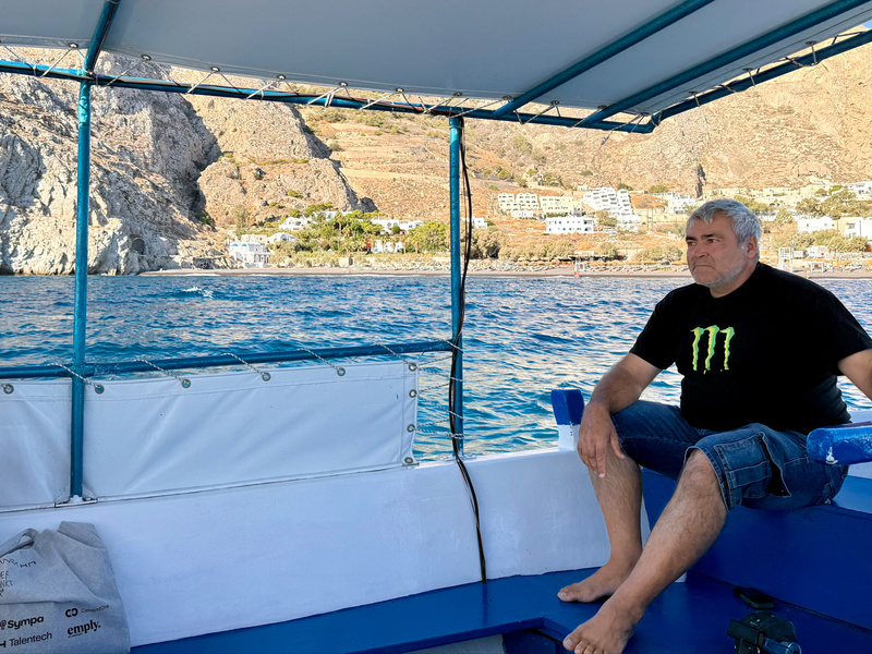 Auto-generated description: A man with gray hair is sitting on a boat, wearing a black shirt and denim shorts, with a view of rocky cliffs and buildings in the background.