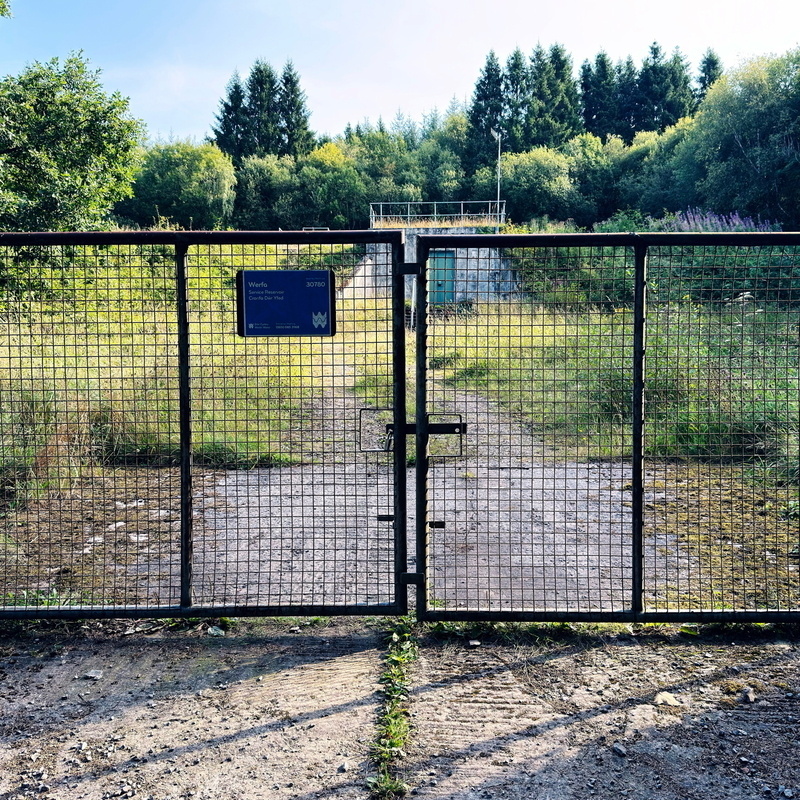 A metal mesh gate in front of an abandoned, overgrown facility surrounded by trees. The pathway inside is weathered, and there’s a sign with some text on the gate. Tall grass and wild plants are visible beyond the gate.