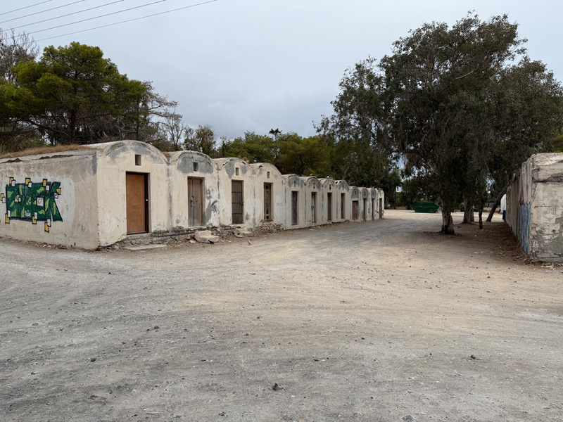 Auto-generated description: A row of abandoned, weathered, concrete buildings with wooden doors sits on a deserted dirt road surrounded by sparse trees.
