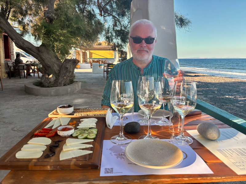 Auto-generated description: A person is sitting at a seaside table with a selection of cheeses, bread, and wine glasses.
