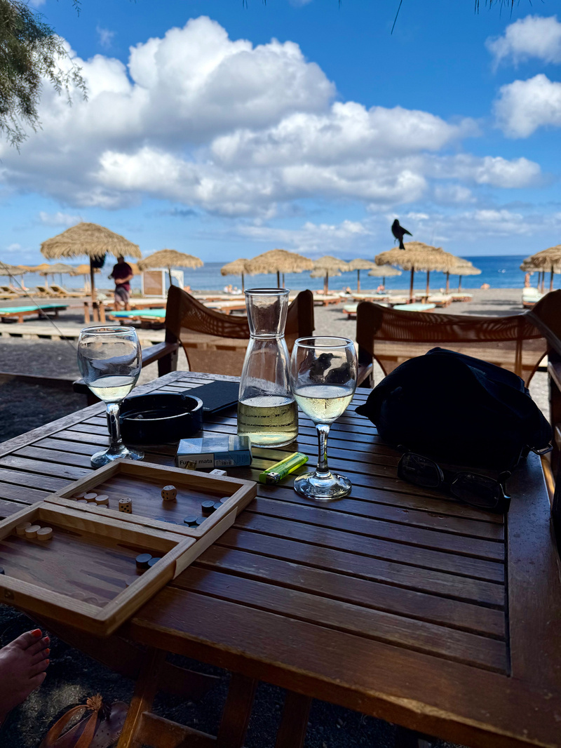 Auto-generated description: A wooden table at a beach setting features wine glasses, a carafe, a backgammon board, and sun loungers with thatched umbrellas in the background.