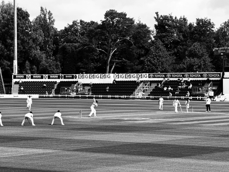 Auto-generated description: A cricket match is in progress with players on the field and an empty stand in the background.