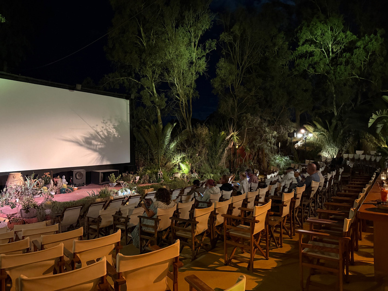 Auto-generated description: An outdoor cinema setup is surrounded by trees, with rows of empty chairs and some people seated, facing a large screen at night.