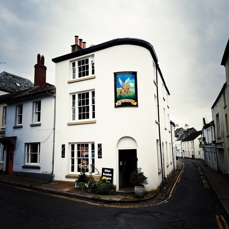 Auto-generated description: A quaint street features a white building with a sign displaying a bird, located in a village setting.