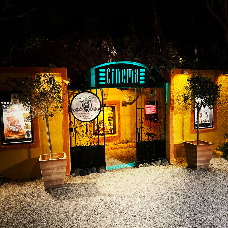 Auto-generated description: A warmly illuminated entrance to a small, rustic cinema with poster displays and potted plants on either side.