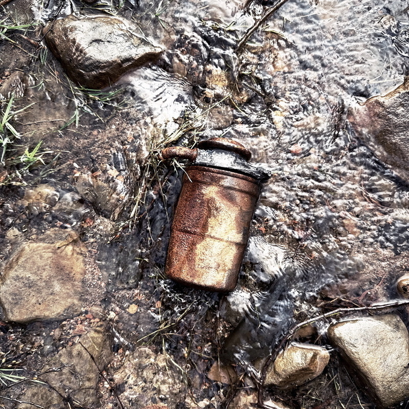A rusted, cylindrical metal object lies on wet, rocky ground surrounded by clear water.
