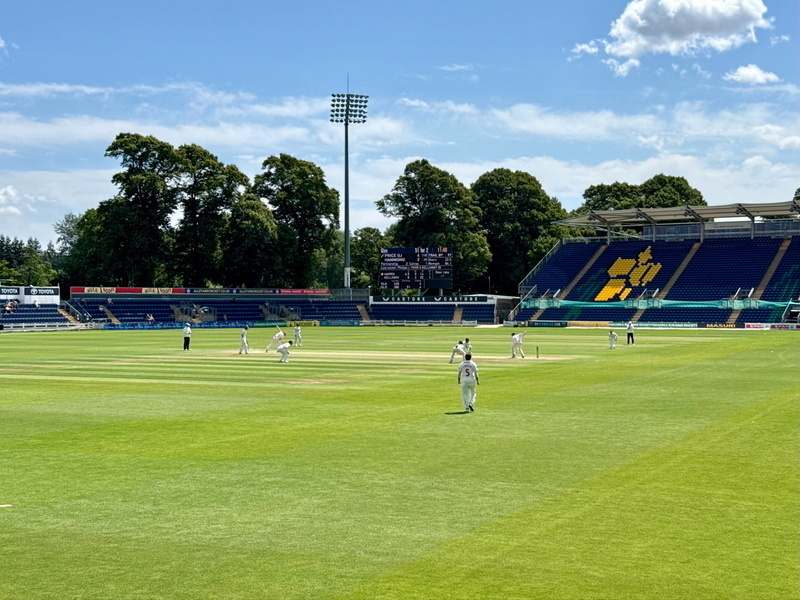 Auto-generated description: A cricket match is taking place in a stadium with a few spectators in the stands under a clear blue sky.