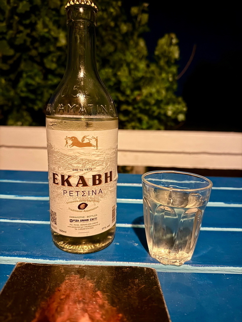 Auto-generated description: A bottle of EKABH Retsina and a filled glass sit on a blue table against a dark background with greenery.