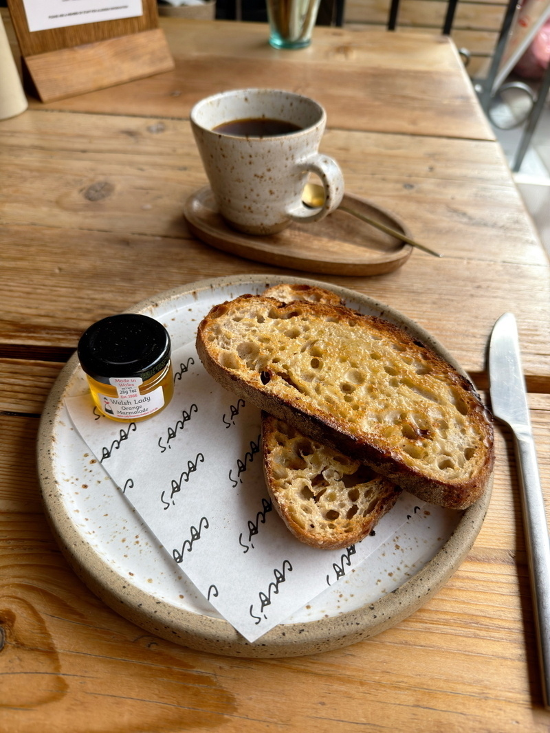 Auto-generated description: A breakfast scene with buttered toast, a small jar of jam, a cup of coffee, and a knife set on a wooden table.