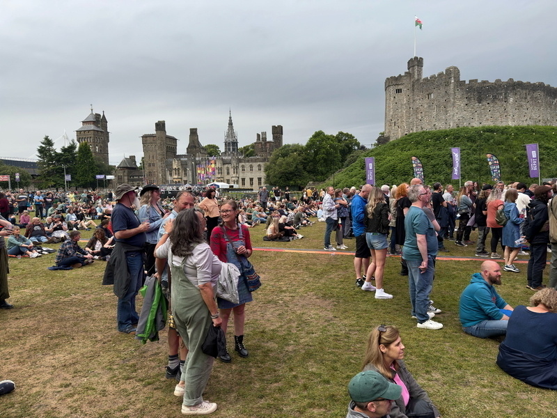 Auto-generated description: A crowd of people is gathered on a lawn near an old stone castle, enjoying an outdoor event.