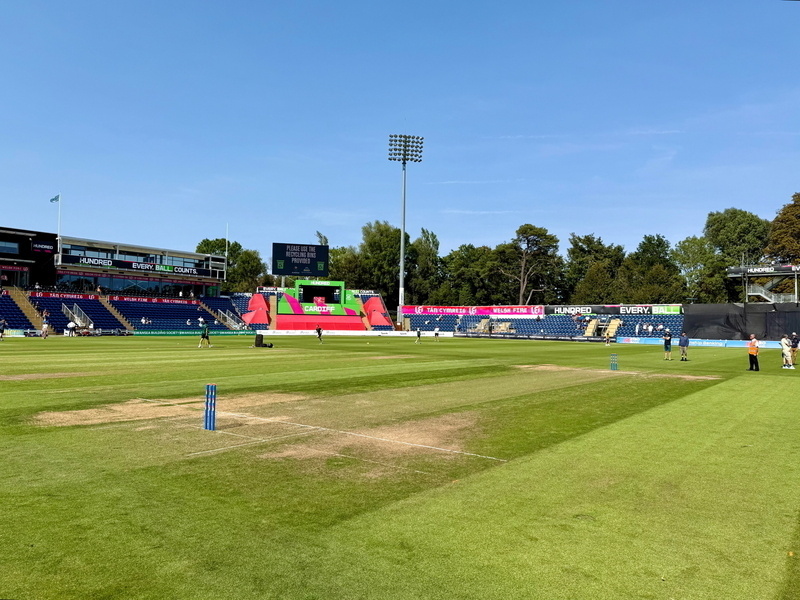 Auto-generated description: A well-maintained cricket ground with markings on the field, surrounded by stands and scoreboards, under clear blue skies.