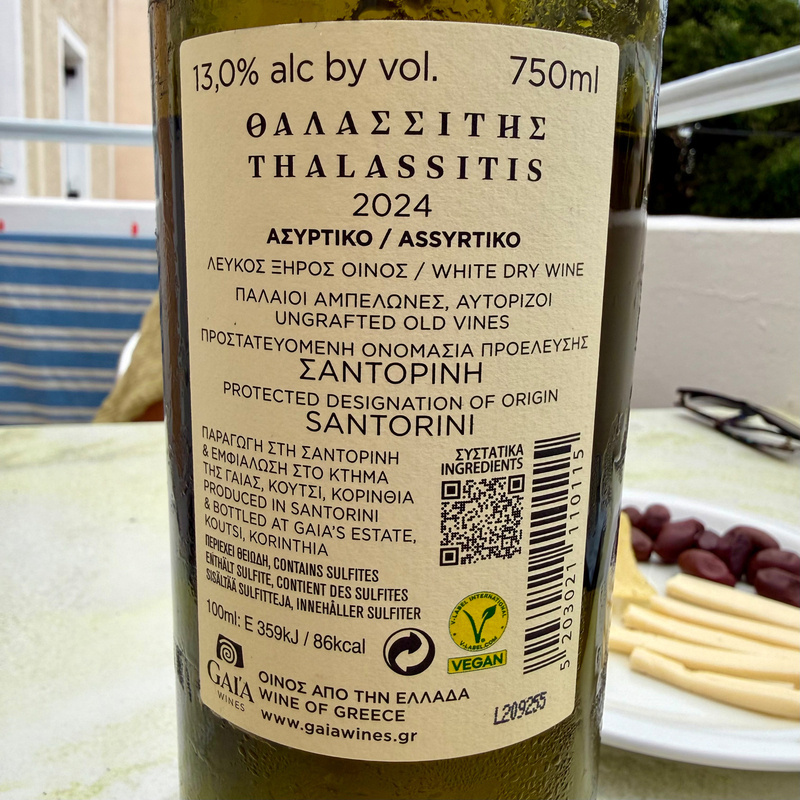 Auto-generated description: A bottle of Thalassitis white dry wine sits on a table with cheese and olives in the background.