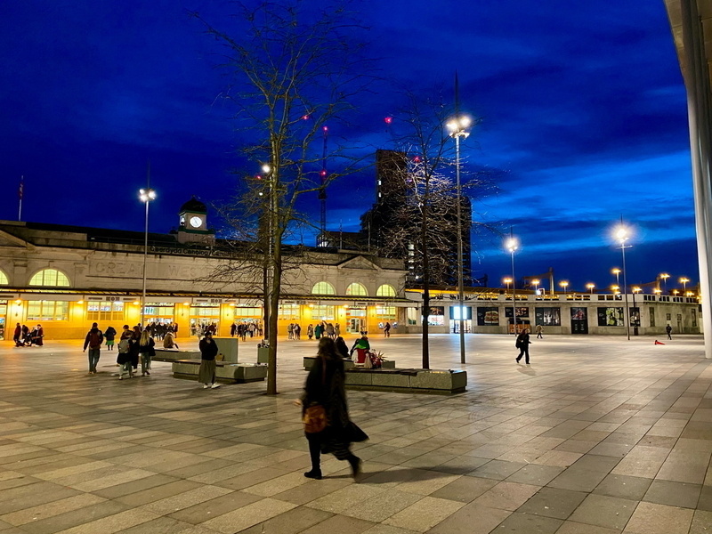 Auto-generated description: A brightly lit urban plaza with people walking, set against a deep blue evening sky and a historic building featuring a clock.