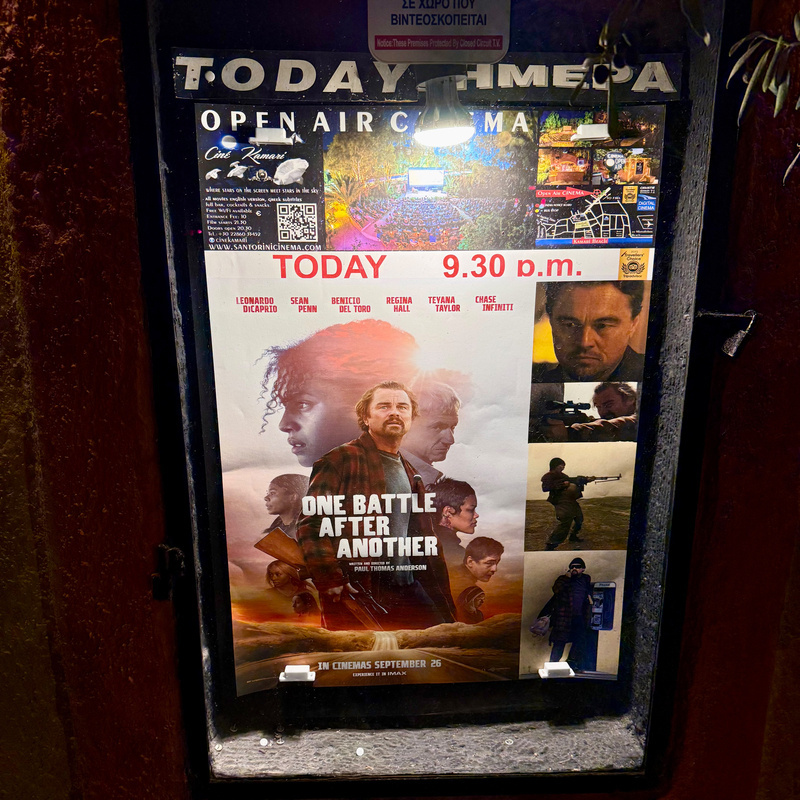 Auto-generated description: A movie poster displayed in an illuminated outdoor cinema showcase advertises the film One Battle After Another with a collage of character images and information about the screening time.