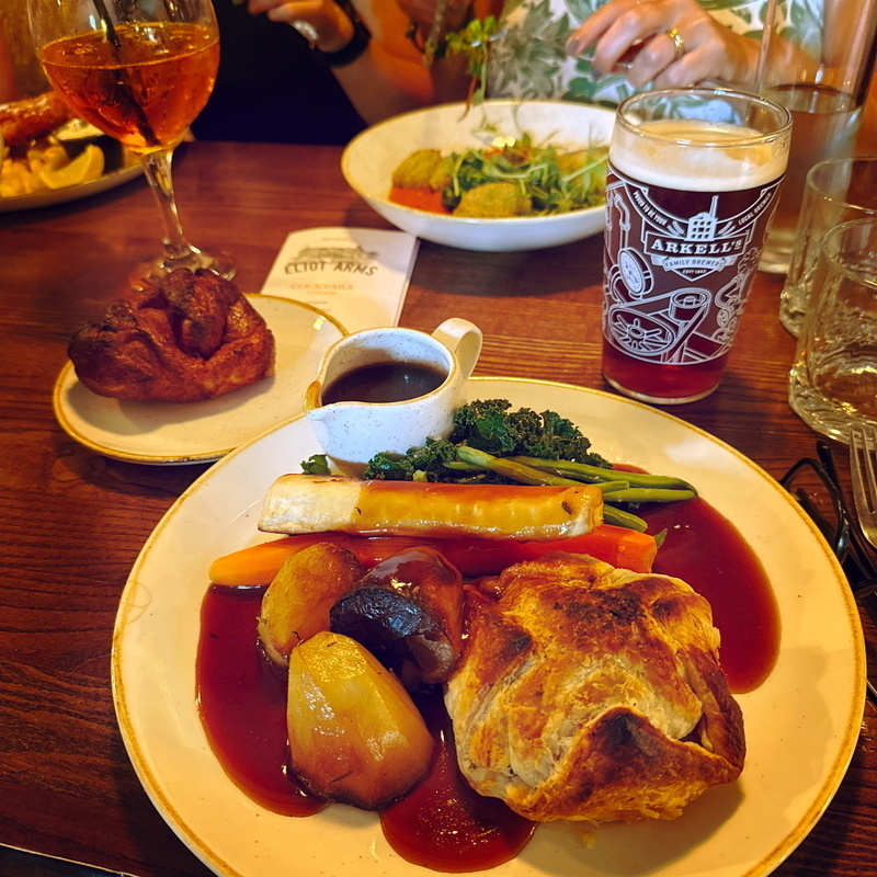 A table with a plate of roast dinner featuring a meat pie, roasted potatoes, carrots, broccoli, and gravy. A Yorkshire pudding is on a separate plate. There's a glass of Aperol Spritz and a pint of beer from Arkell's Brewery.