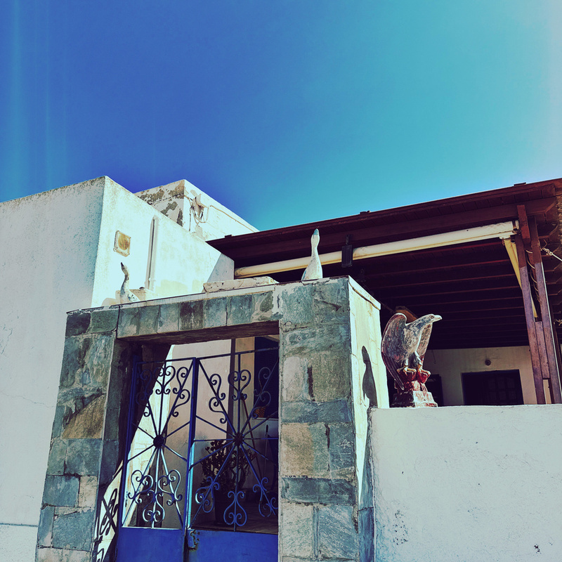 A rustic stone gate leads to a house with blue doors and a rooftop adorned with decorative elements under a clear blue sky.