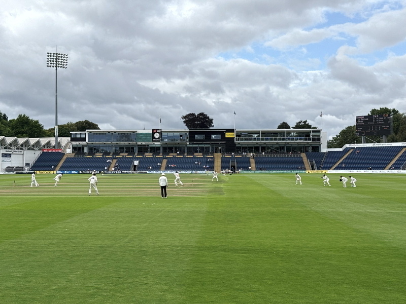 Auto-generated description: A cricket match is being played in a stadium with players positioned around the field and a large seating area in the background.