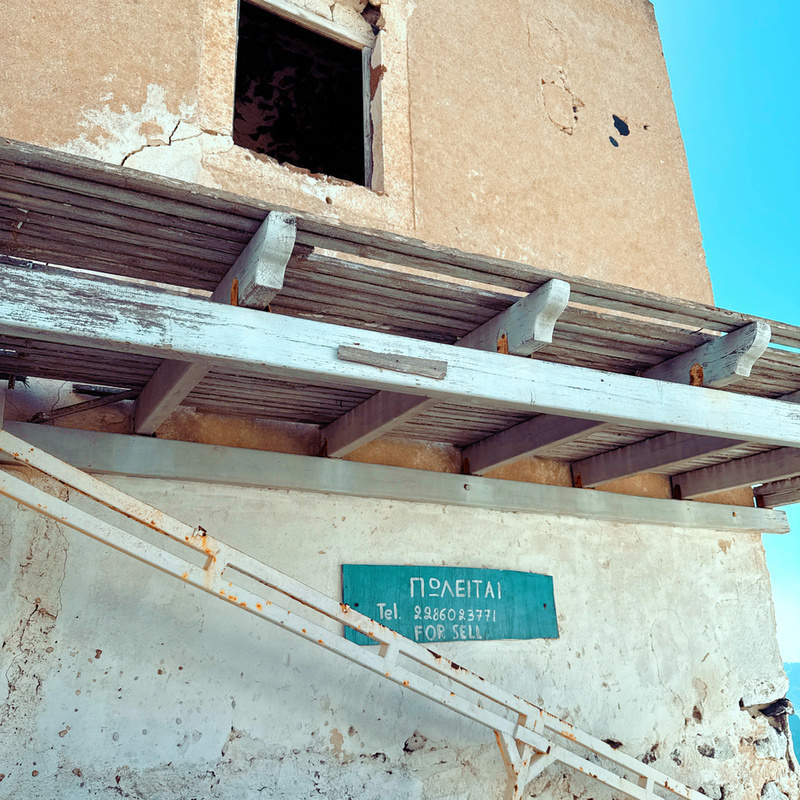 A weathered building with a partially open window and a sign that reads ΠΩΛΕΙΤΑΙ in Greek, indicating it's for sale.