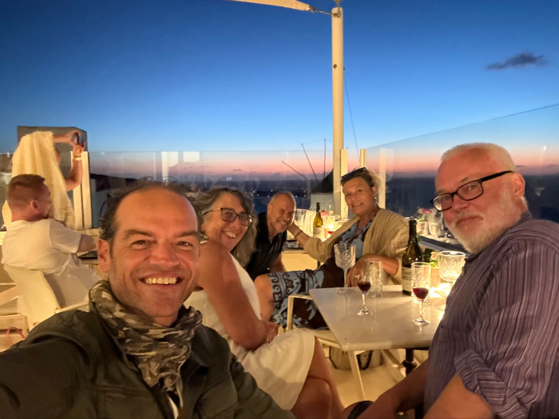 Auto-generated description: A group of people is sitting together at a table outdoors during sunset, smiling and enjoying drinks.
